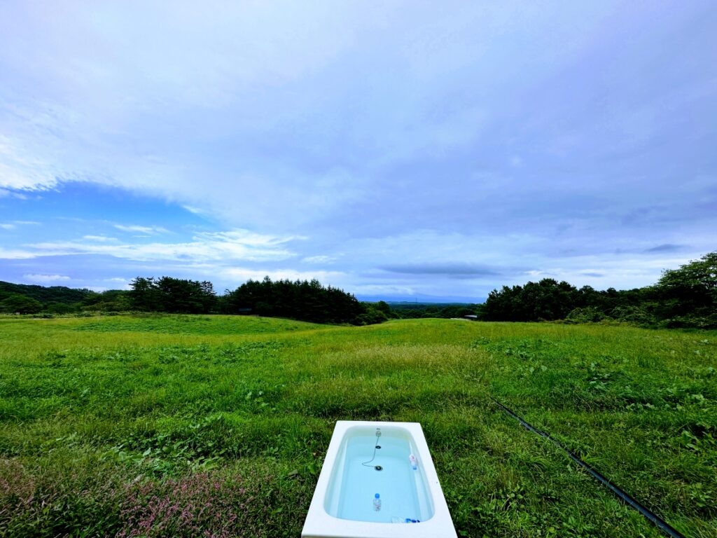 那須の広大な緑の丘に置かれた白いバスタブの水風呂。地下50mから汲み上げた天然水が注がれ、背景には抜けるような空が広がる。