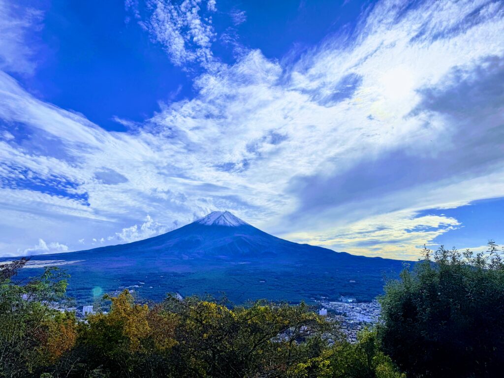 富士山パノラマロープウェイ山頂付近からの眺望。青空と筋雲を背景に、雪化粧した富士山の全景と、その麓に広がる河口湖周辺の街並みが一望できる絶景パノラマ。