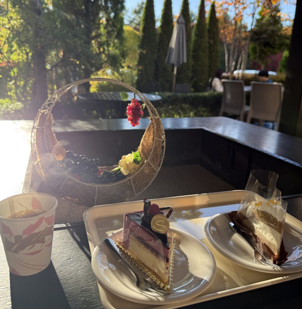 河口湖チーズケーキガーデンのテラス席。白地に金色のロゴが入ったケーキ2種類と、淹れたてのコーヒーがテーブルに並び、背景には富士山麓の豊かな緑が広がっている。