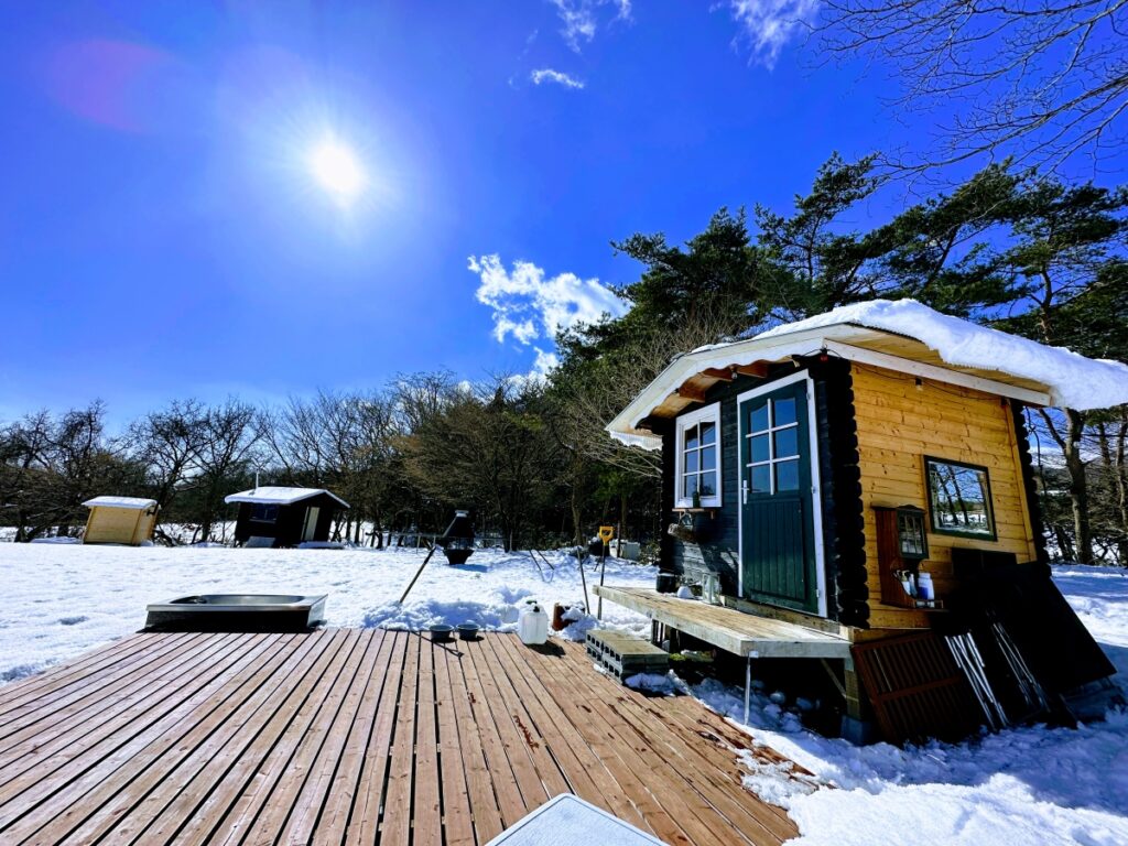 栃木県那須町のアウトドアサウナ「Quiet Storm Nasu」の冬景色。青空と一面の雪原に佇むサウナ小屋とウッドデッキの風景。