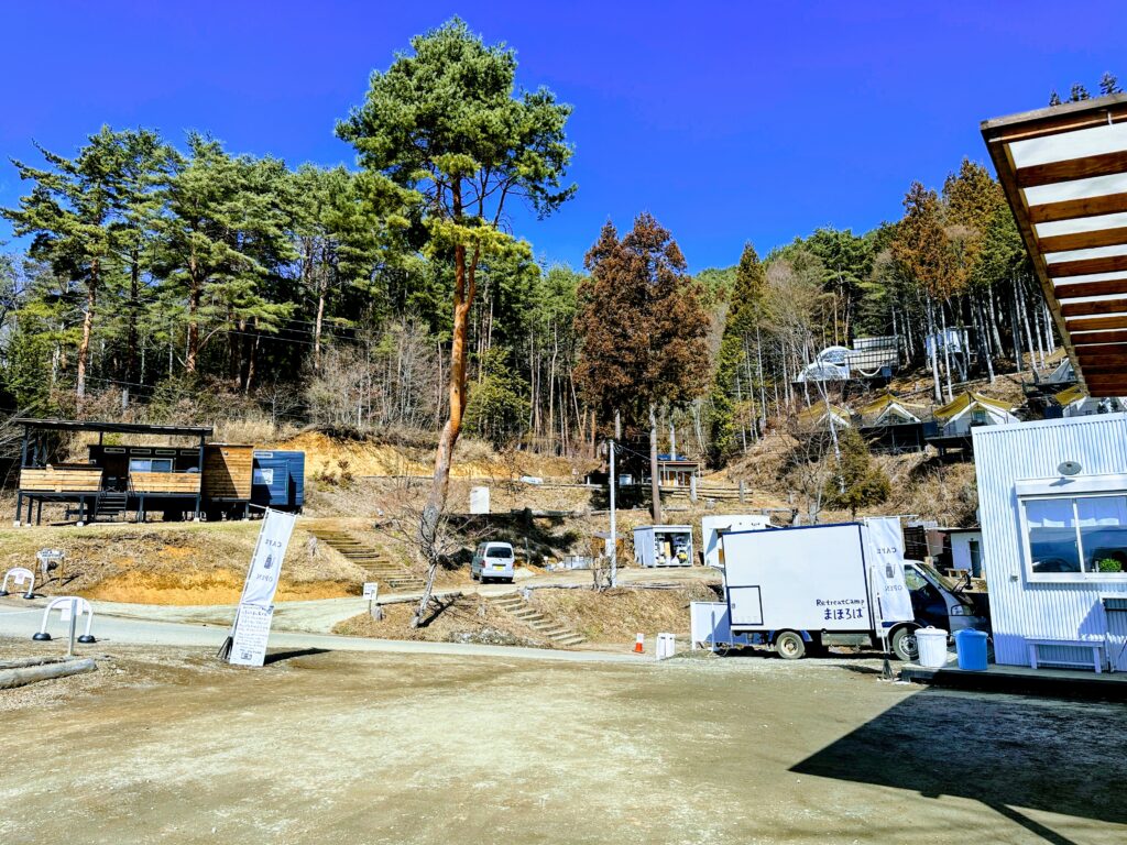 青空の下、河口湖を見下ろす高台に広がる「Retreatcamp まほろば」のキャンプ場全景。看板のある入り口付近と、斜面に並ぶ宿泊コテージ。
