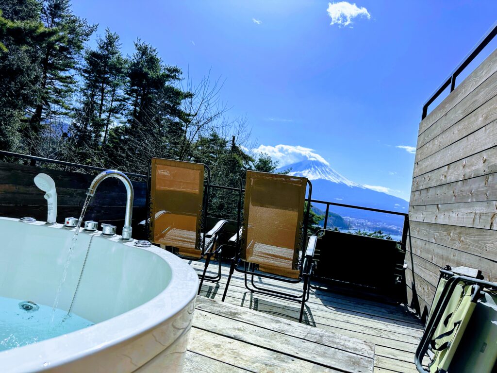 Retreatcamp まほろばの展望デッキ。手前に水が流れる白亜のジャグジー、中央に富士山を正面に望む2台のインフィニティチェア、背景には青空と雪化粧した富士山、河口湖が広がる絶景。
