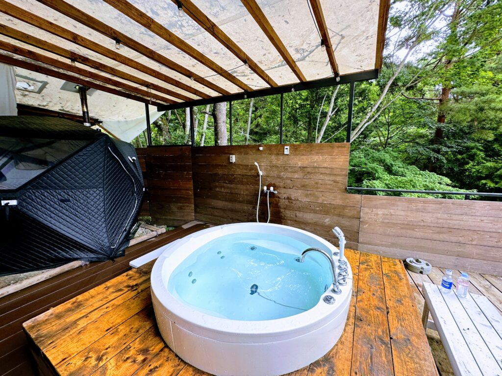 Retreatcamp まほろばのサウナデッキ中央に設置された、真っ白な円形ジャグジー。左手には黒いテントサウナ（MORZH）があり、周囲は豊かな木々に囲まれている。