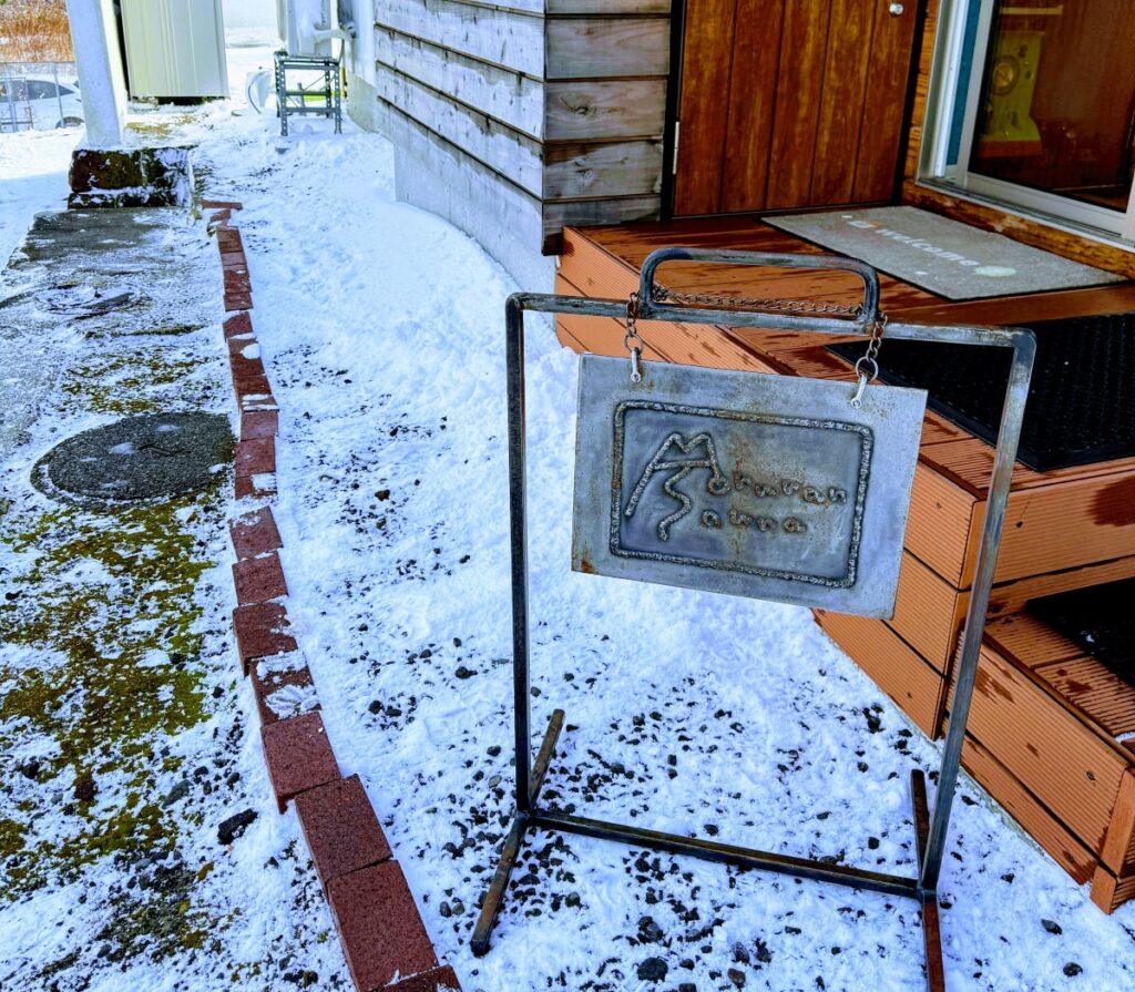 積雪の中に設置された、「Moruran Sauna」の文字が刻まれたアンティーク調のアイアン看板。背景には木造のサウナ小屋の入り口が見える。