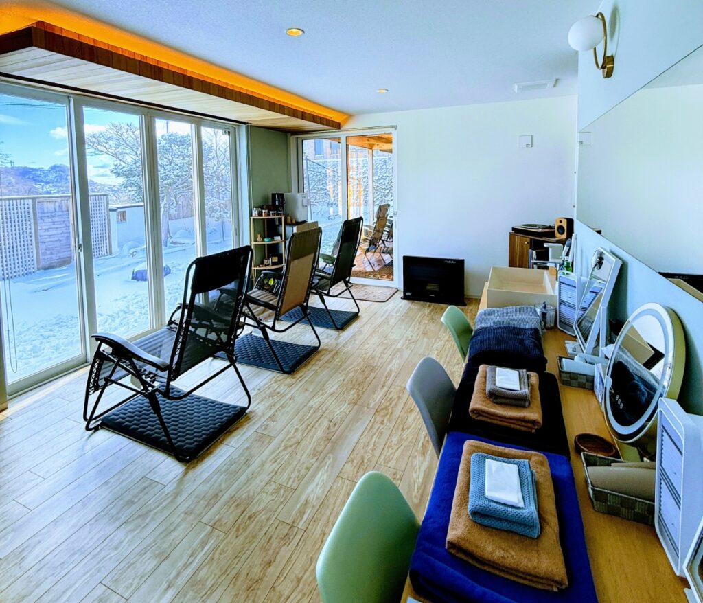 大きな窓から雪の積もった庭の景色が一望でき、室内には複数のインフィニティチェアが並んでいる。右側のカウンターにはタオルやスキンケア用品、ドライヤーなどのアメニティが整然と用意されている。