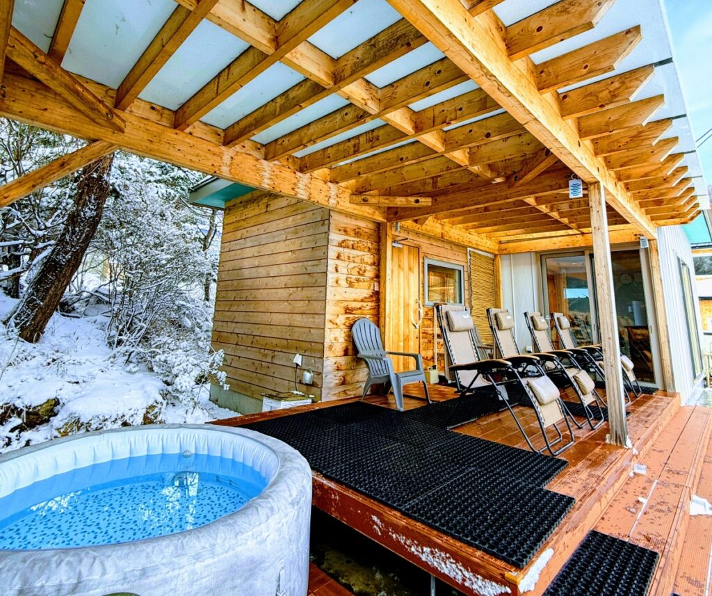 雪が積もった屋外テラスに設置された、円形のジャグジー付き水風呂。テラスにはアディロンダックチェアやリクライニングチェアが並び、雪景色が広がっている。