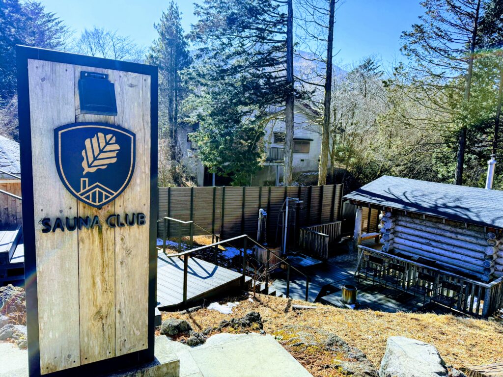 サーマルクライムスタジオ富士の屋外エリア「The Mountain」の入り口にある、ロゴマークが刻まれた木製の看板と奥に見えるケロサウナ。