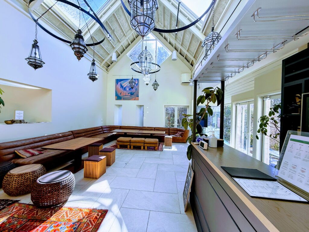 サーマルクライムスタジオ富士のラウンジ「The Lounge」。高い吹き抜けの天井と天窓から差し込む明るい陽光、壁沿いに配置された長いブラウンのソファ、異国情緒あふれる吊り下げランタン、そして清潔感のある白いタイル床が広がる開放的な空間。