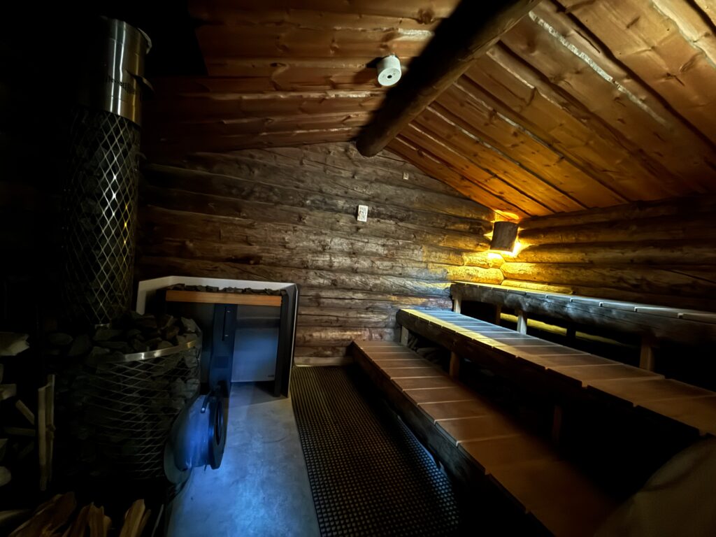 サーマルクライムスタジオ富士の屋外エリア「The Mountain」にあるケロサウナ内部。中央に鎮座する巨大な薪ストーブと、希少な「ケロ材」を使用した重厚なログ壁、温かみのある間接照明。