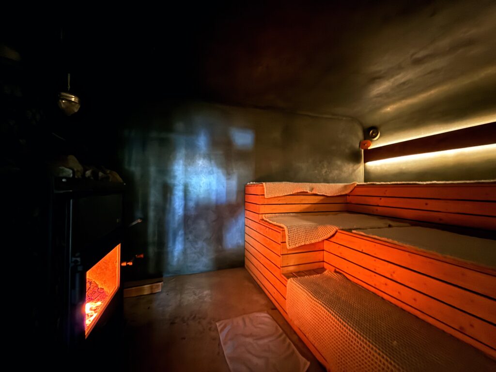 「第2章：五感を揺さぶる「薪ストーブ」の魔力。82度から100度へ進化させた熱の正体」という見出しと説明文が付いた、NAYA SAUNA（ナヤサウナ）のサウナ室内部の写真。左側に炎が燃える巨大な薪ストーブ、右側に3段構成の木製座面があり、間接照明が落ち着いた雰囲気を演出している。