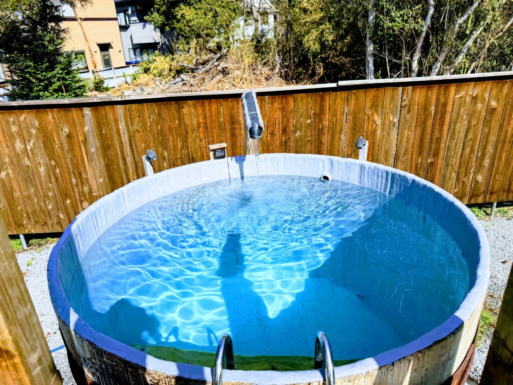 青空の下に設置されたNAYA SAUNAの屋外水風呂。木枠の大きな樽風呂に澄み切った地下水が注がれ、周囲は木製の柵で囲まれている。