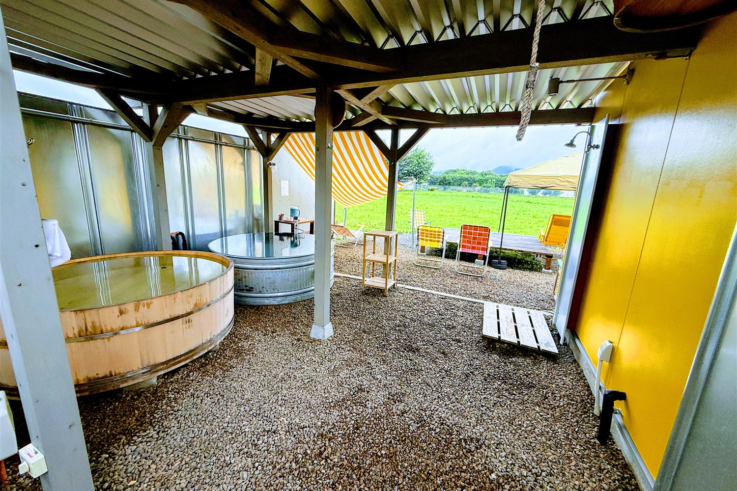 WALLOWS sauna & dinerの屋根付きスペースに設置された金物の水風呂タンクと木桶の風呂、奥に広がる原っぱ