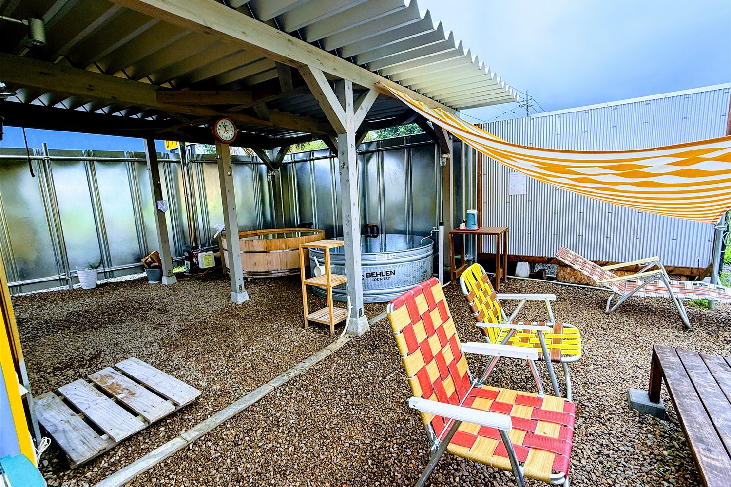 WALLOWS sauna & dinerの休憩スペース。レトロなガーデンチェアが並び、屋根とタープの下に水風呂が設置されている