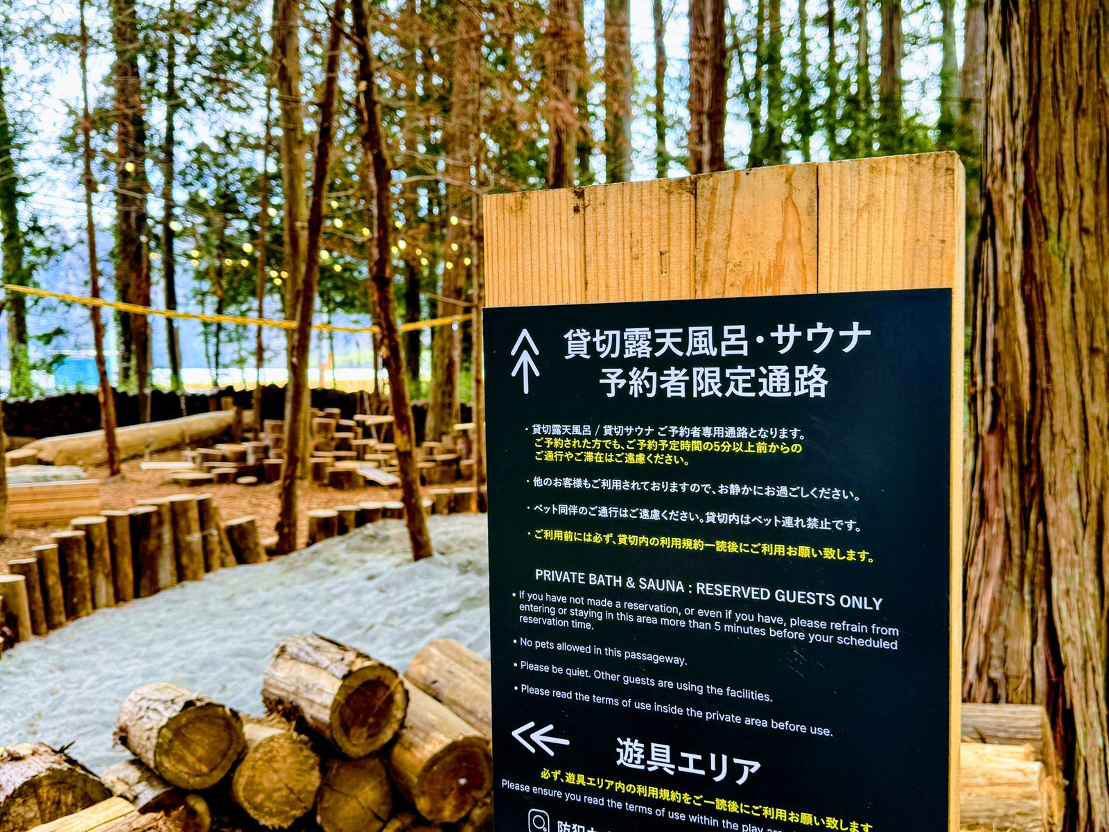 Render Fika Sammuの貸切露天風呂・サウナ予約者限定通路の案内看板と、積まれた薪、木立