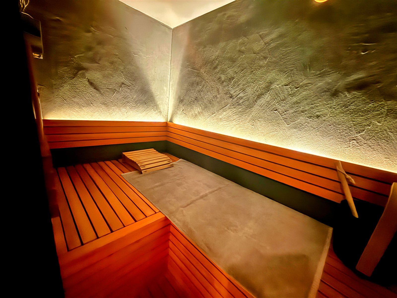 PRIVATE SAUNA maa & GROCERYのサウナ室内部。モルタル調の壁に間接照明、木のベンチとサウナマット、ロウリュ用のバケツとラドル