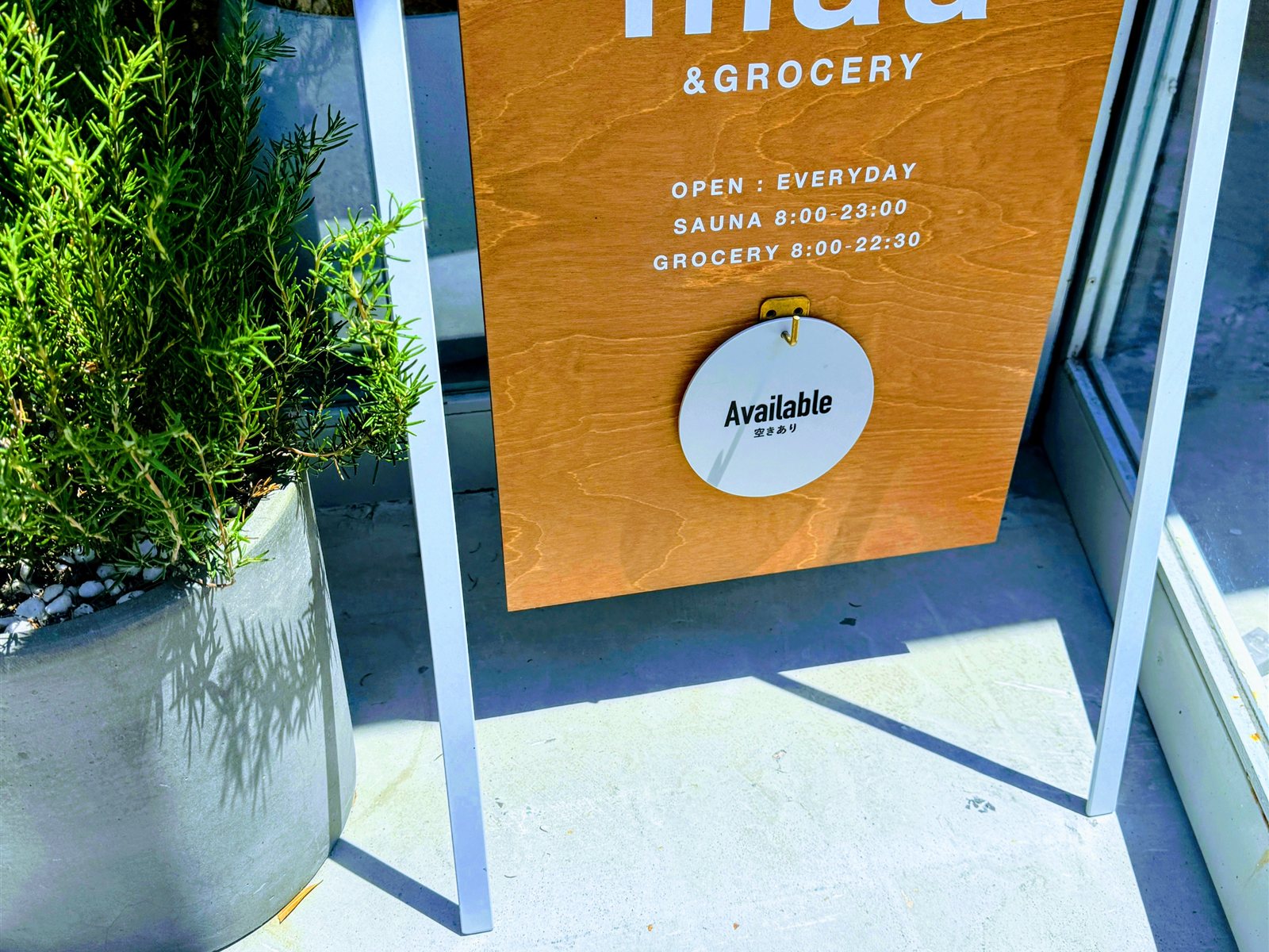 PRIVATE SAUNA maa & GROCERYの木製A型看板。OPEN EVERYDAY、SAUNA 8:00-23:00、GROCERY 8:00-22:30、Available（空きあり）の表示