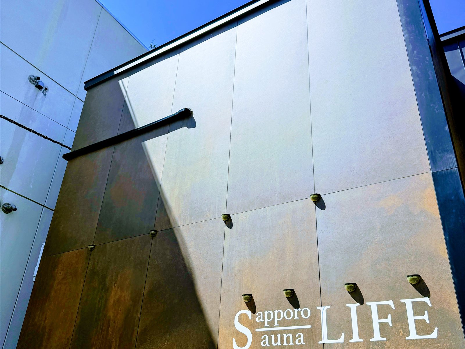 PRIVATE SAUNA S-LIFEの外観。ブロンズ調のメタリックな壁面に「Sapporo Sauna LIFE」のロゴ、青空が広がる