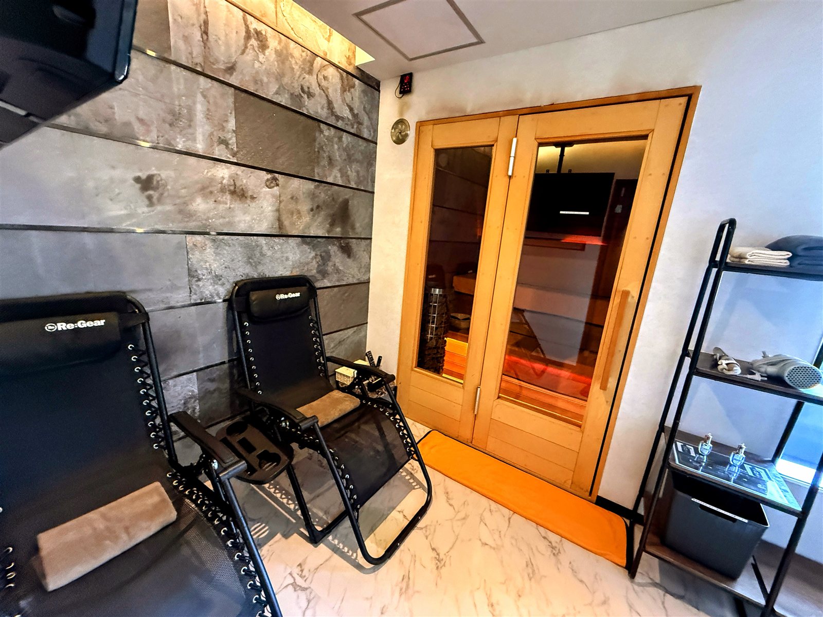 PRIVATE SAUNA S-LIFEの個室全体。ガラス扉のサウナ室、インフィニティチェア、ストーン調の壁とテレビが一室にまとまっている