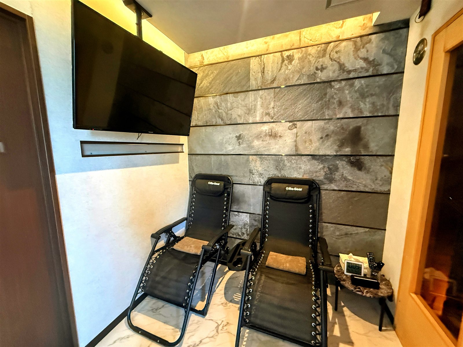 PRIVATE SAUNA S-LIFEの休憩スペース。Re:Gearのインフィニティチェアが2台、壁掛けテレビ、ストーン調の壁