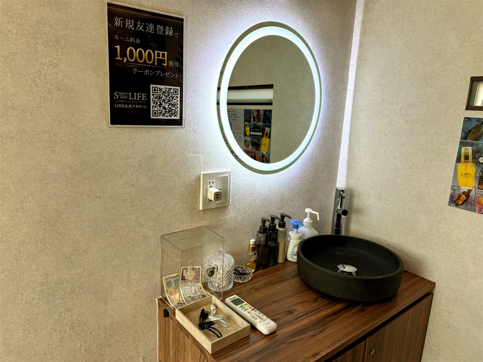 PRIVATE SAUNA S-LIFEのパウダースペース。LED付き丸鏡、黒い石の洗面ボウル、アメニティ類、LINE公式アカウントの案内ポスター
