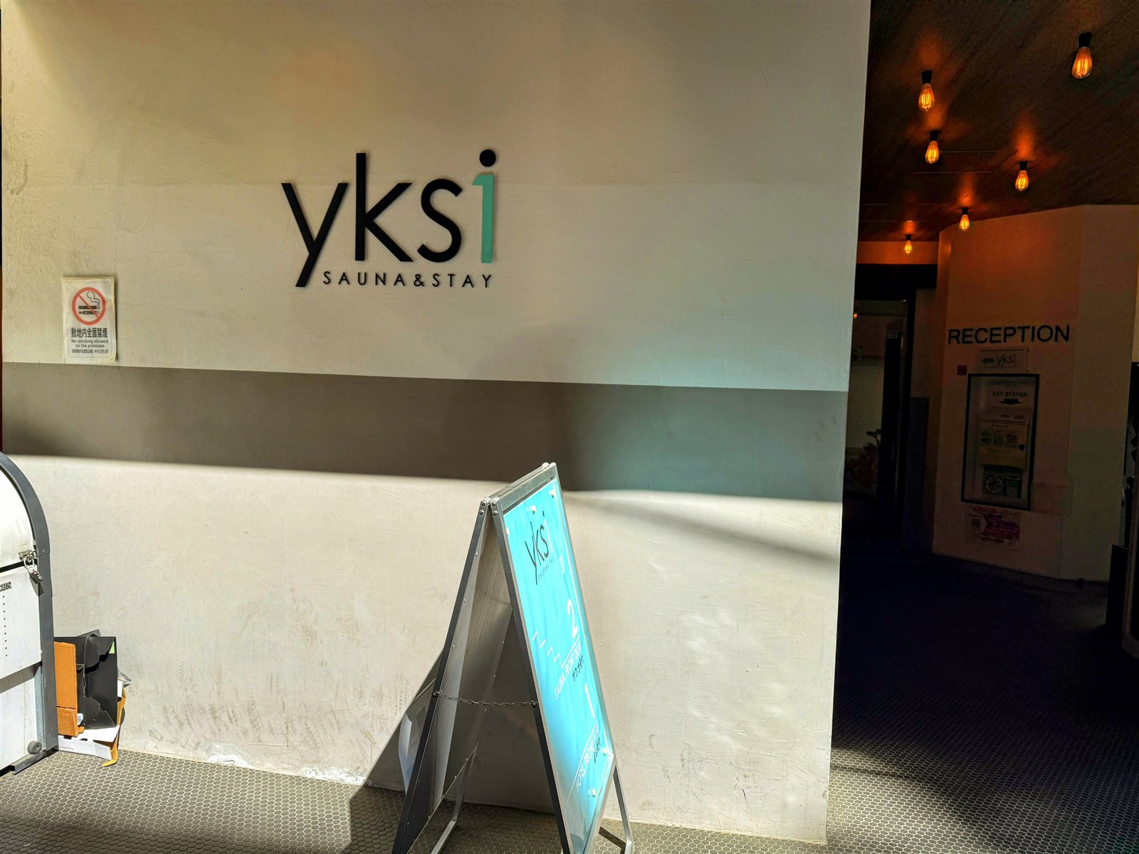 yksi SAUNA & STAYの入口。白い壁にyksi SAUNA&STAYのロゴと、奥にRECEPTIONの案内が見える