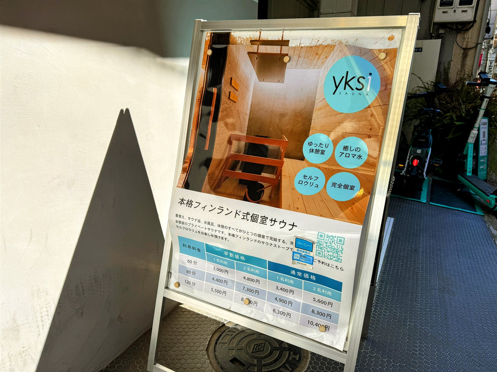 yksi SAUNA & STAYの案内看板。本格フィンランド式個室サウナの特徴（ゆったり休憩室・癒しのアロマ水・セルフロウリュ・完全個室）と料金表が掲示されている