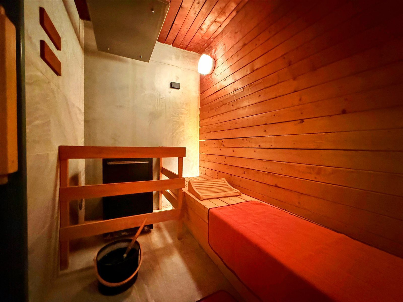 yksi SAUNA & STAYの個室サウナ室内部。木のベンチに赤いマットと木製の枕、足元にロウリュ用のバケツとラドルが置かれている