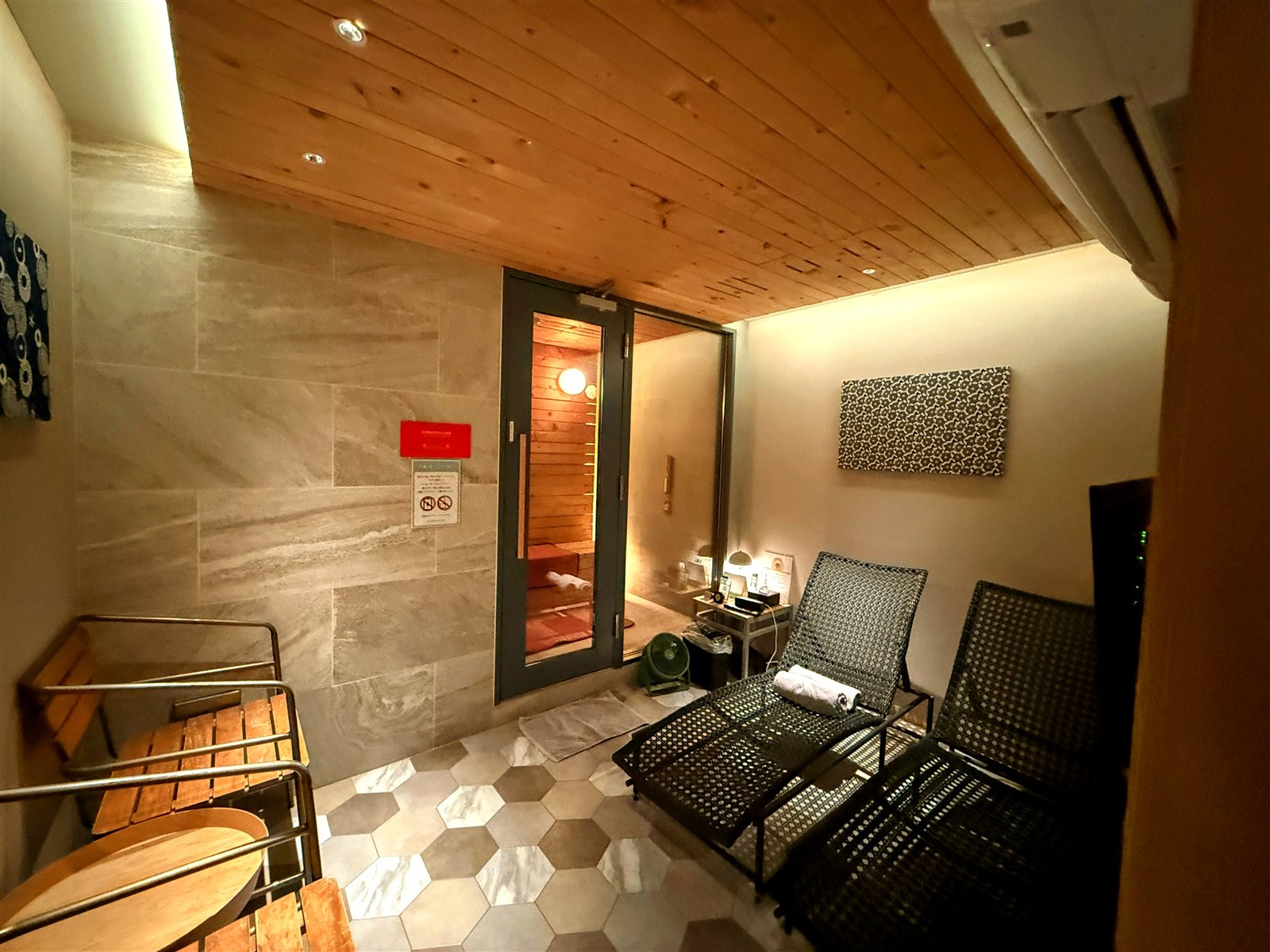 yksi SAUNA & STAYの個室内の休憩スペース。黒いデッキチェアが2脚並び、ガラスドアの奥にサウナ室が見える。床はヘキサゴンタイル
