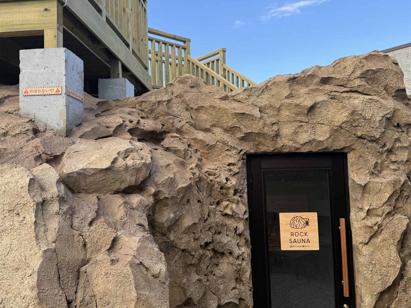 YUBUNE SAUNA PARKのロックサウナの入口。ゴツゴツした岩肌の壁に「ROCK SAUNA」の木製サイン、青空が広がる