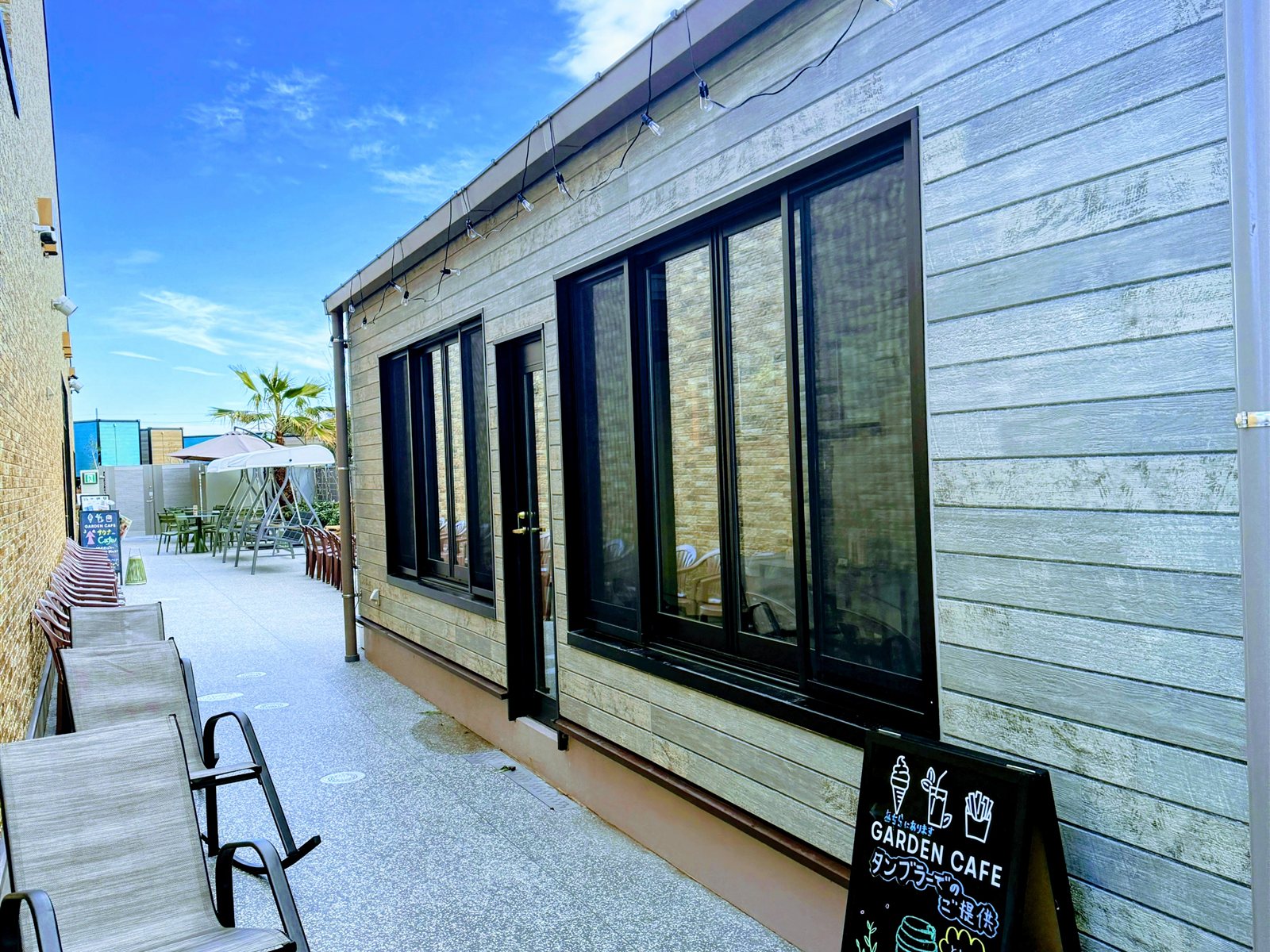 YUBUNE SAUNA PARKの休憩スペース。建物沿いにデッキチェアが並び、奥にGARDEN CAFEの案内と椰子の木、青空