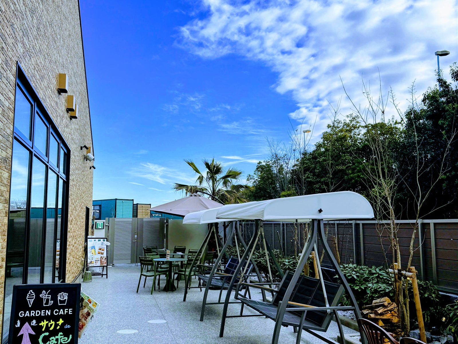 YUBUNE SAUNA PARKのガーデンエリア。屋根付きのブランコベンチやガーデンチェアが並び、椰子の木と青空、GARDEN CAFEの案内