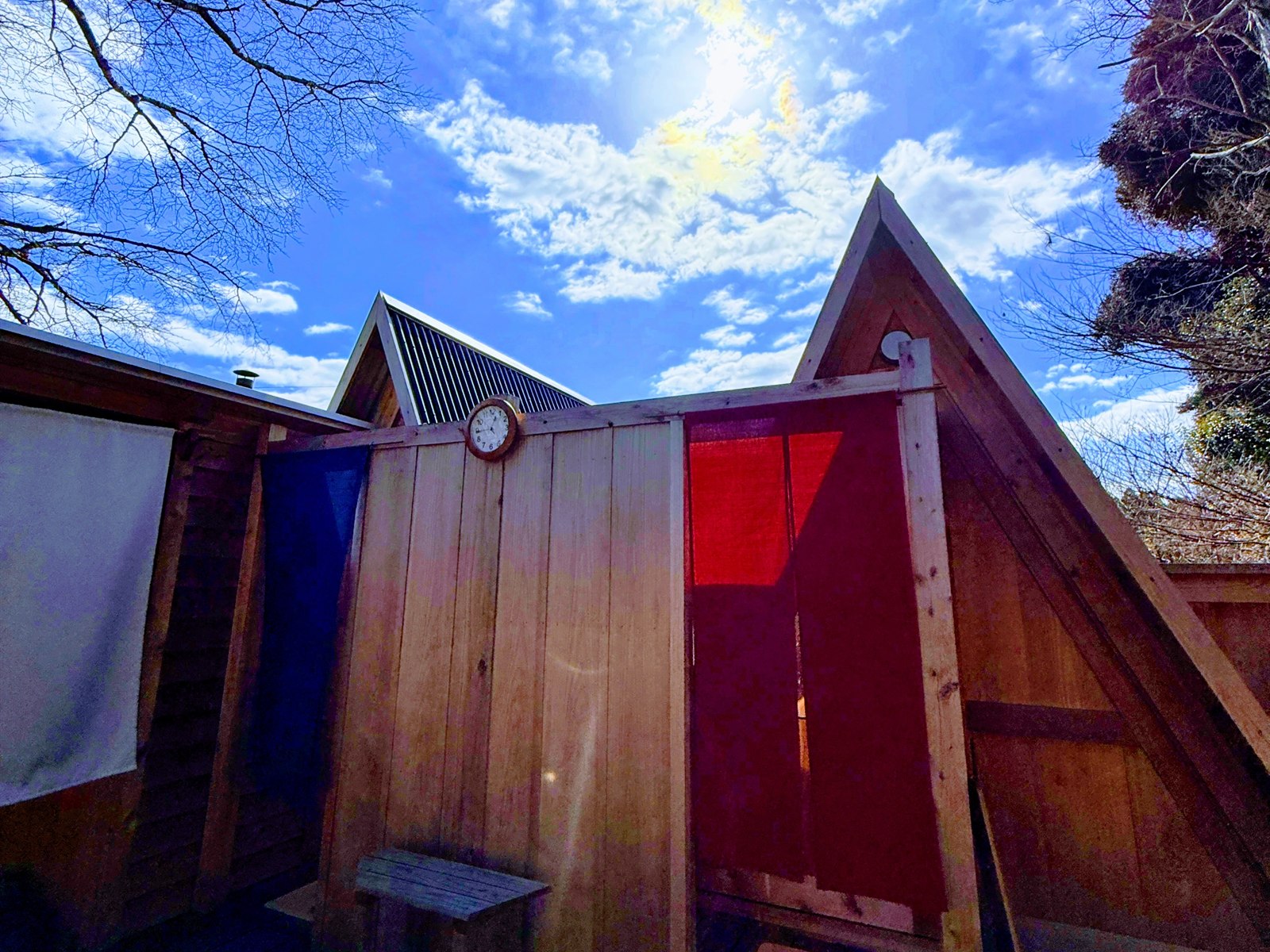 小江戸サウナの更衣室。三角屋根の木の小屋と青と赤ののれん、青空